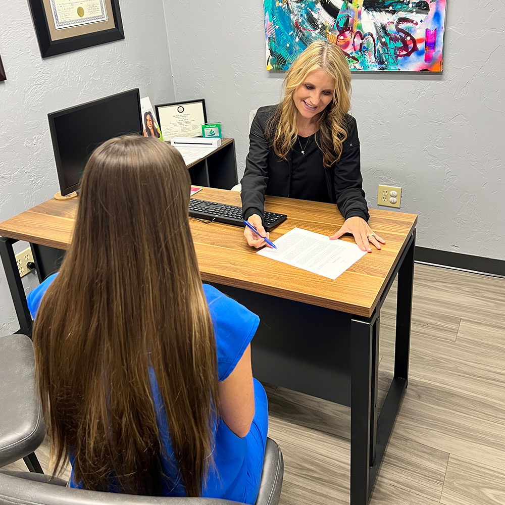 The image shows a woman sitting at a desk with a pen and paper, engaged in conversation with another person who is standing and holding a piece of paper.