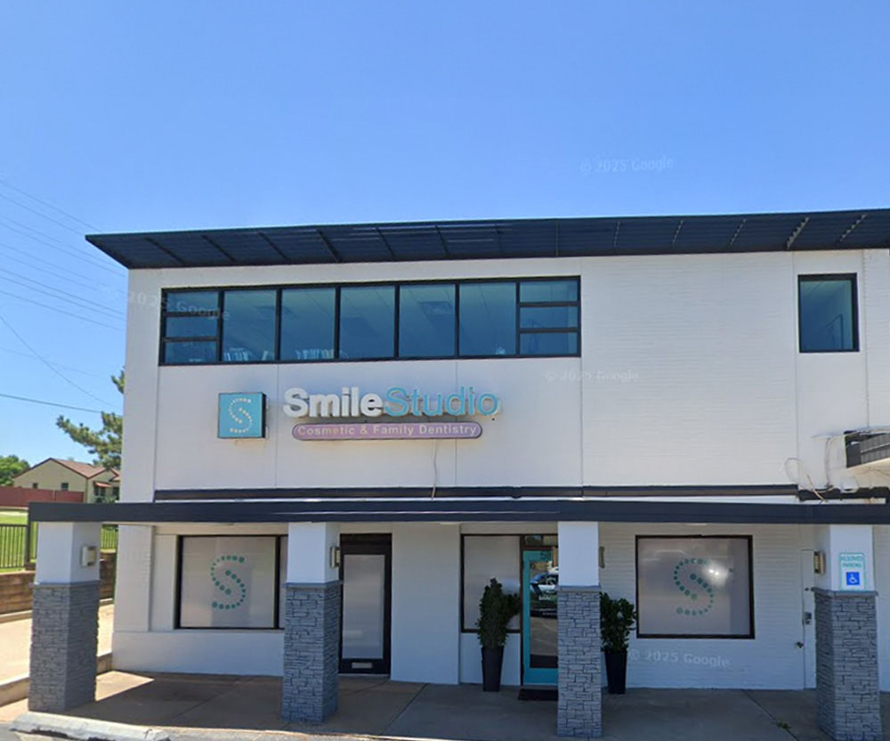 The image shows a modern two-story building with a white exterior, featuring a large storefront window on the ground floor and a smaller one above it. The signage includes SMILE STUDIO prominently displayed in blue letters at the top of the front facade. There is also a logo that appears to be a smiley face, suggesting a connection to dental services or a similar theme. The building has a flat roof with solar panels and a green awning over the storefront window. The sky is clear and blue, indicating fair weather conditions.