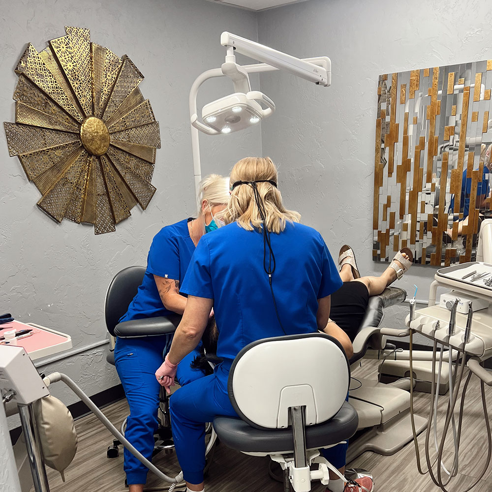 The image depicts a dental office setting where two dental professionals are seated at a chair with dental equipment, working on a patient who is lying down and appears to be receiving dental care.